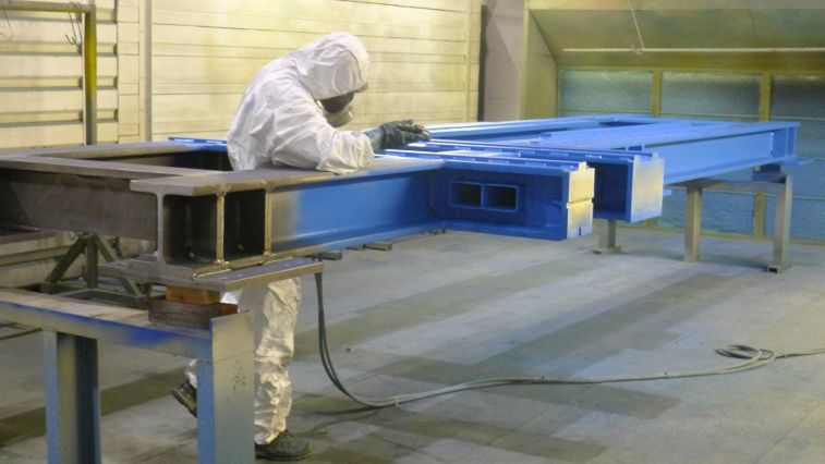 Ein Bauteil wird von einem Mitarbeiter in Schutzkleidung blau lackiert